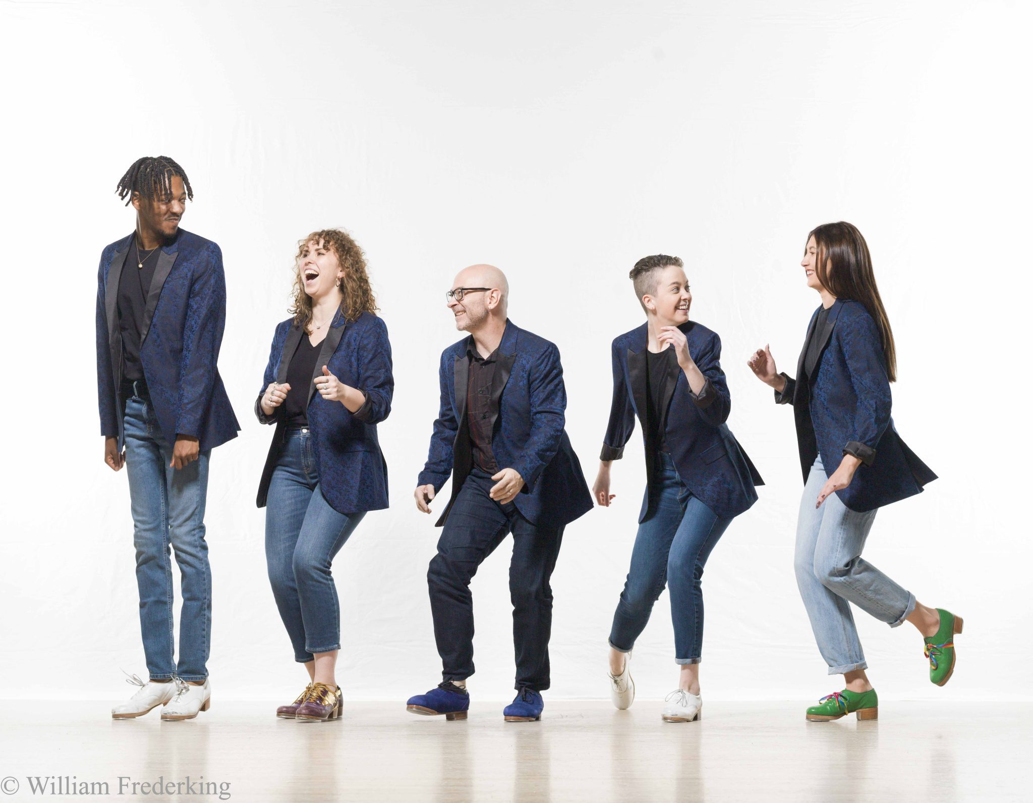 Five people, three women and two men, stand in a line against a plain white background. They wear matching dark blazers and jeans, striking dynamic, playful poses. Each person has a distinct hairstyle and tap shoe footwear, adding individuality to their coordinated outfits.
