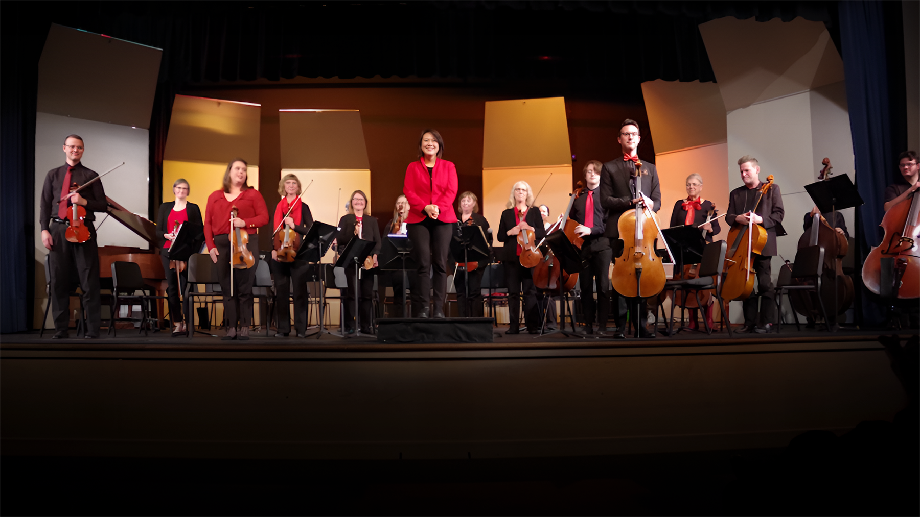 A group of musicians on stage, each holding various string instruments including violins, cellos, and double basses. They are dressed in black and red and appear to be finishing a performance or rehearsal. The background features lit panels in yellow and orange hues.