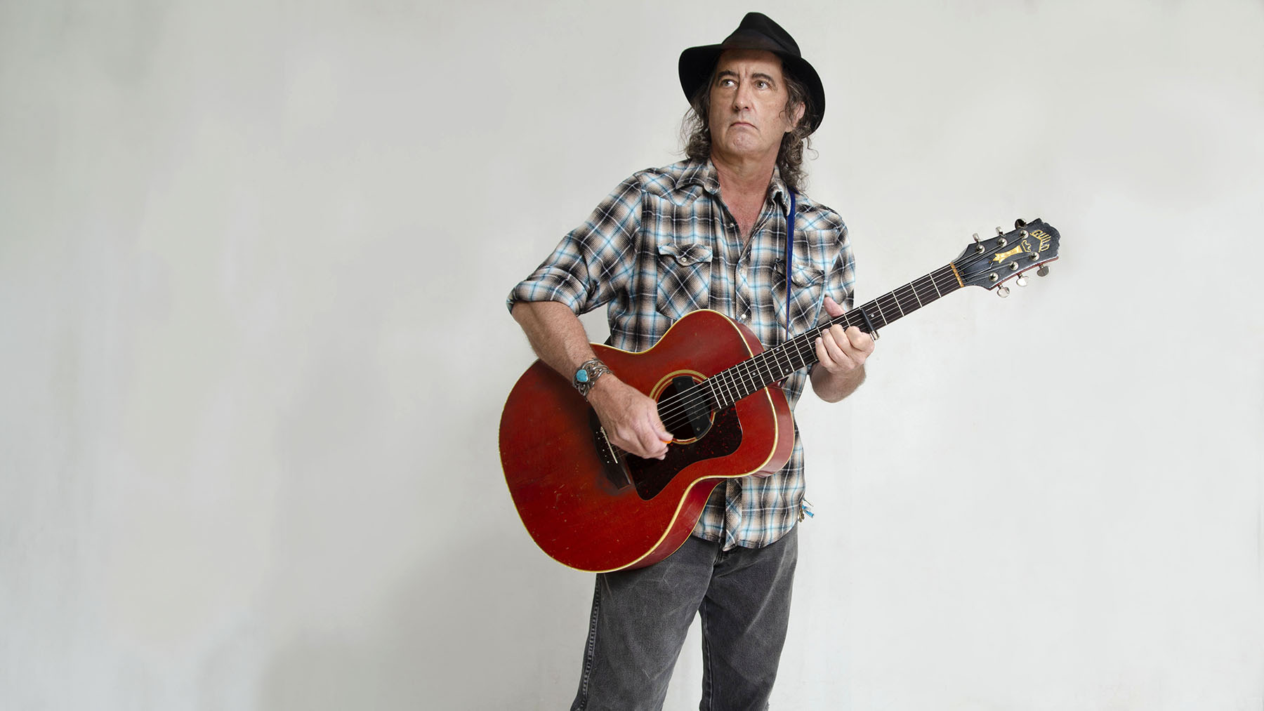 A man with long hair, wearing a black hat, plaid shirt, and jeans, holds and strums a red acoustic guitar against a plain white background, looking into the distance.