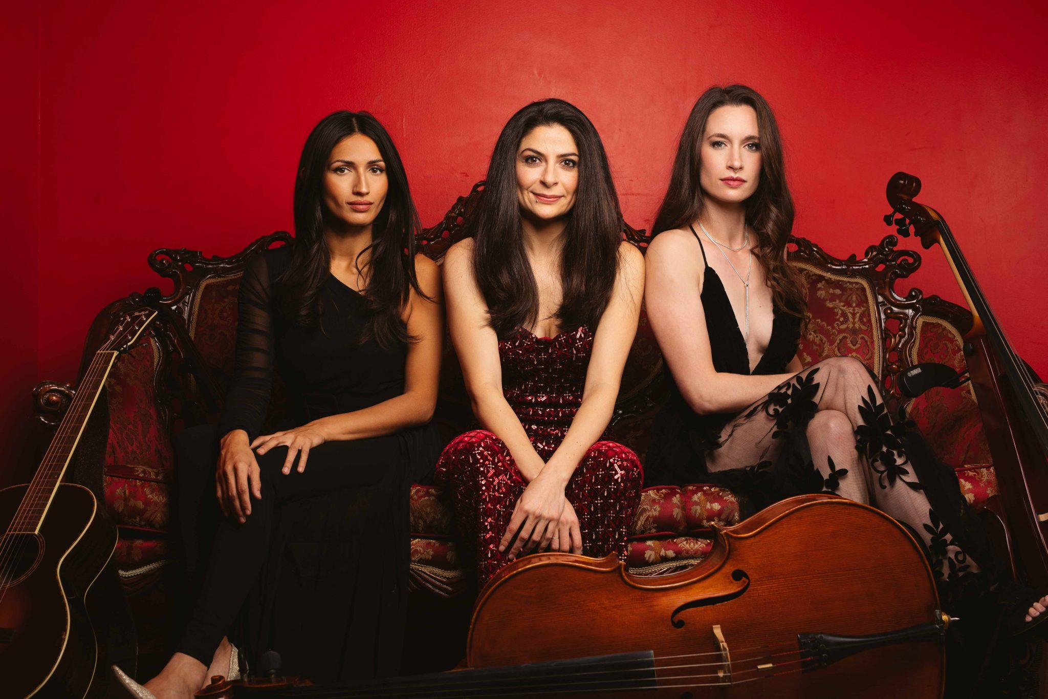 Three women sit on an ornate sofa against a red background, each holding a musical instrument. The woman on the left holds a guitar, the woman in the center has a cello, and the woman on the right has a violin. They wear elegant dresses and pose confidently.