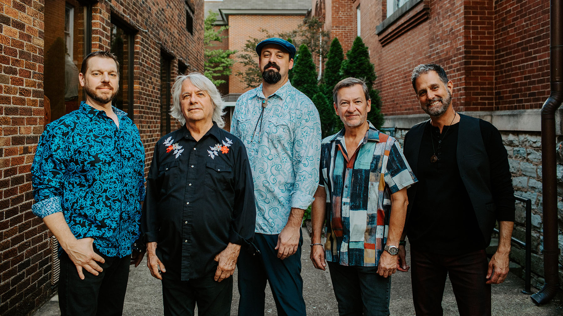 Five men stand in an alleyway between brick buildings. They are dressed in patterned shirts. The group poses casually, with some smiling, against a backdrop of greenery and bricks.