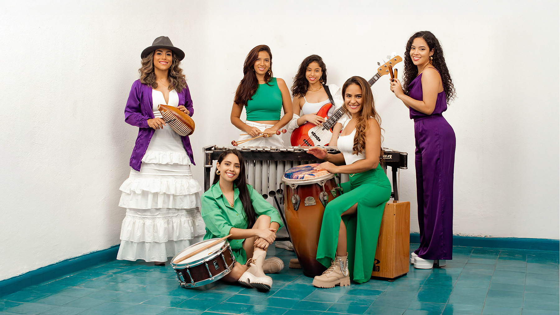 Six women musicians pose together indoors with various instruments, including congas, a güiro, bass guitar, vibraphone, snare drum, and a cajón. They wear colorful, coordinated outfits and smile at the camera.