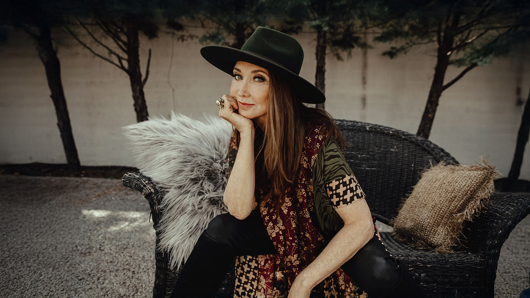 A woman wearing a wide-brimmed dark hat and patterned top sits on a wicker chair with a furry cushion, resting her chin on her hand, outdoors by a white wall with trees in the background.