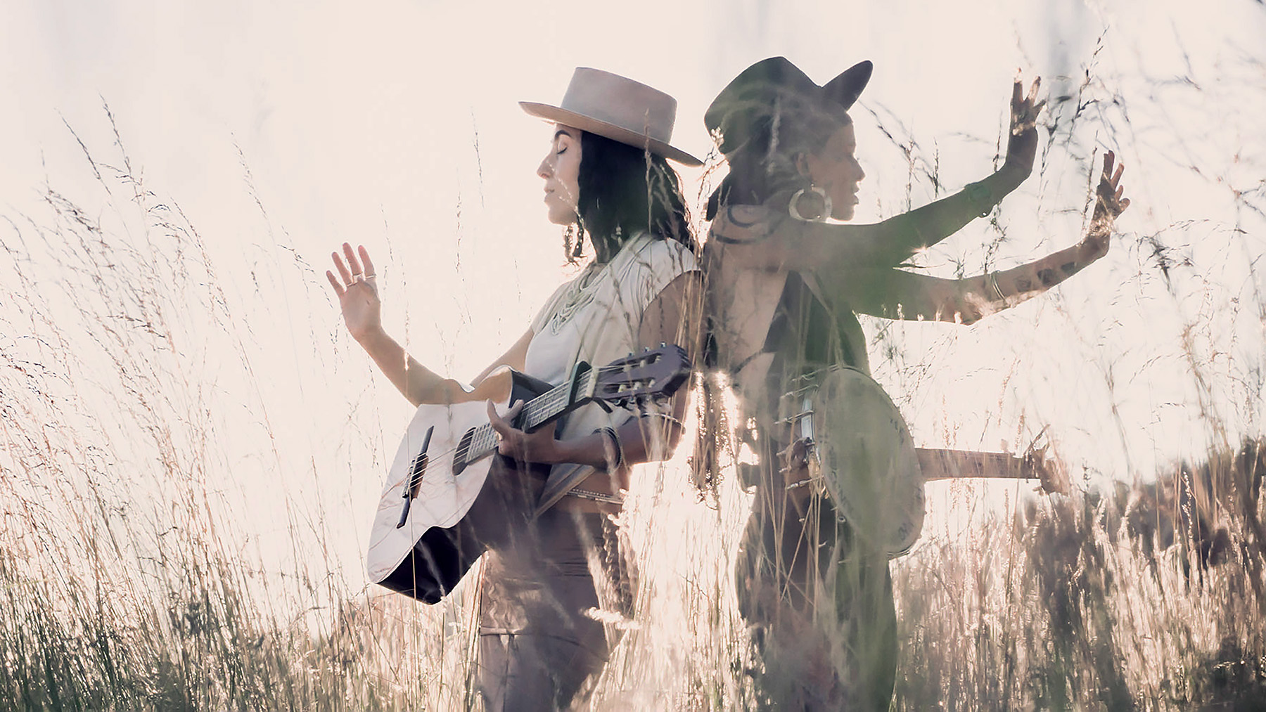 Two women stand back to back in a sunlit field, one holding a guitar. The photo is artistically double-exposed, creating layered images of both women with their arms raised, blending with tall grass.