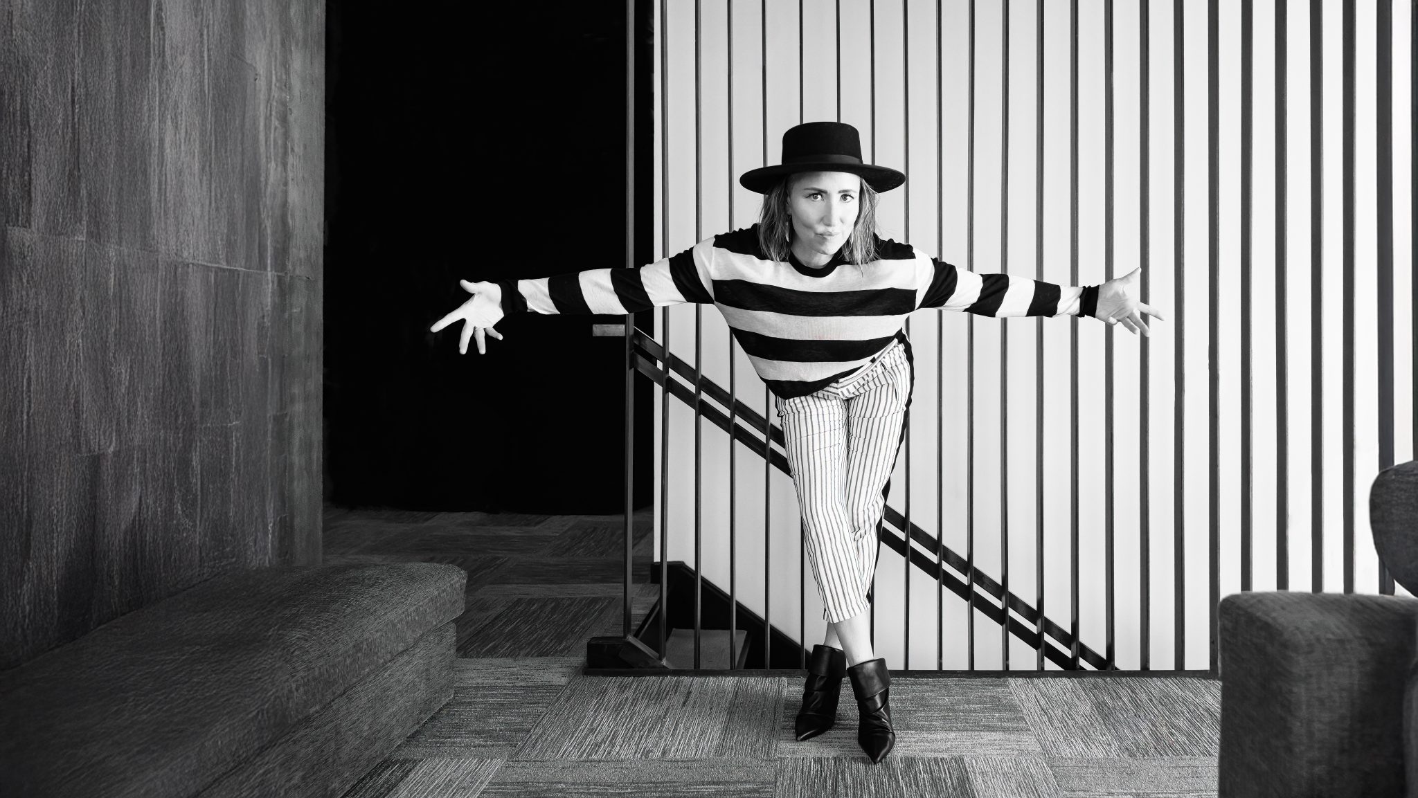 A person in a striped sweater, striped pants, and a wide-brimmed hat poses playfully with arms outstretched in a modern room with vertical lines on one wall. The image is in black and white.